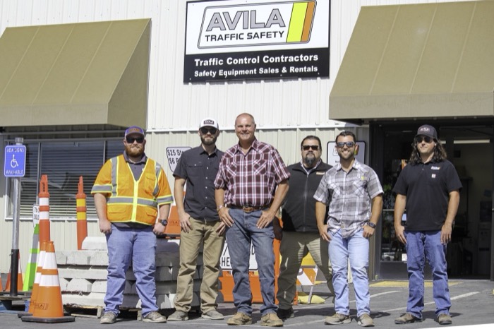 Mike Avila standing with five members of his crew outside the Avila Traffic Safety building, traffic cones stacked nearby