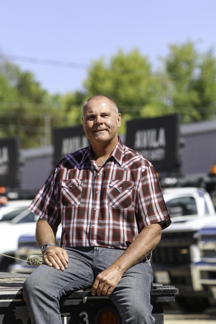 Mike Avila seated on the back of a truck, Avila company trailers parked behind him